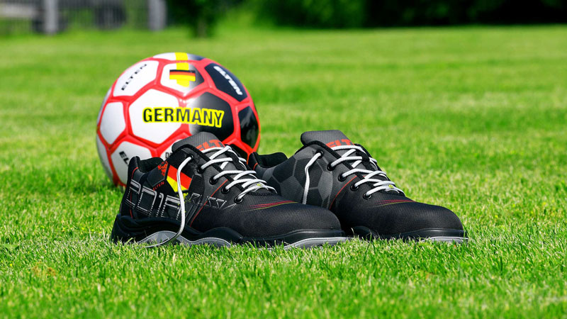  Der Sicherheitsschuh GERMANY Low S3 von Elten vor einem Deutschland Fußball auf grünem Rasen.